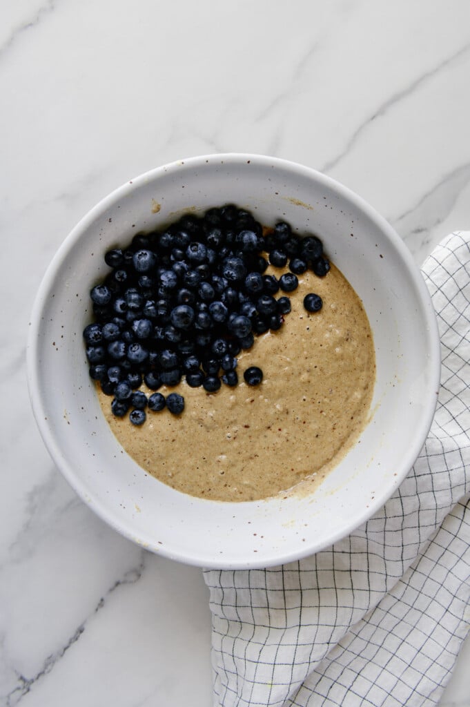 Blueberries added to mixture for vegan blueberry muffins in a white mixing bowl.