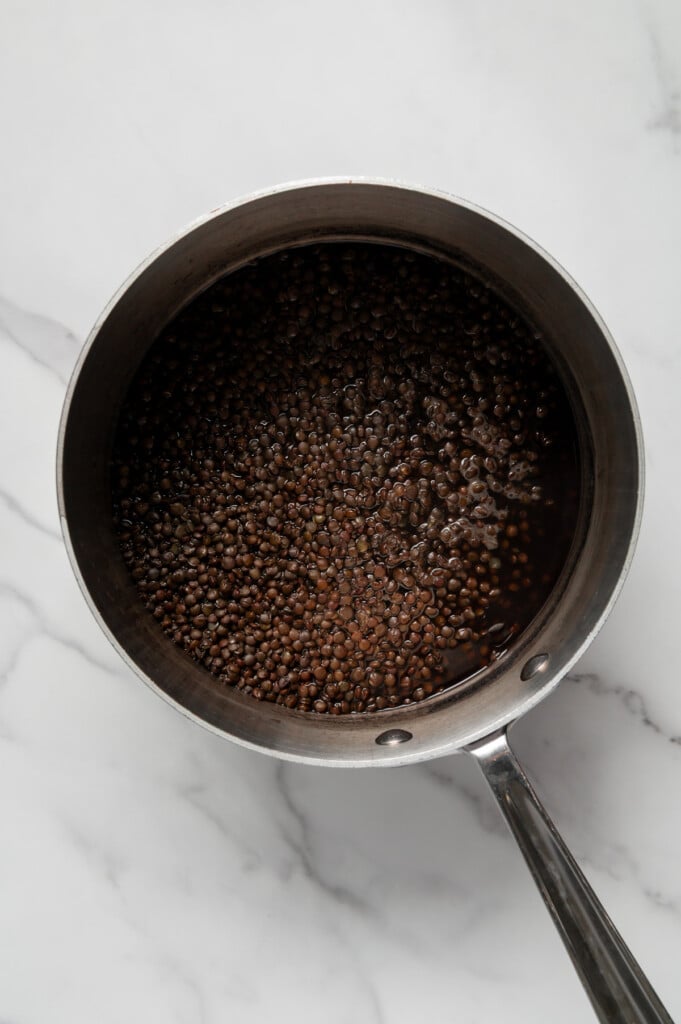 Black lentils cooking in water in a sauce pan with a white marble background.