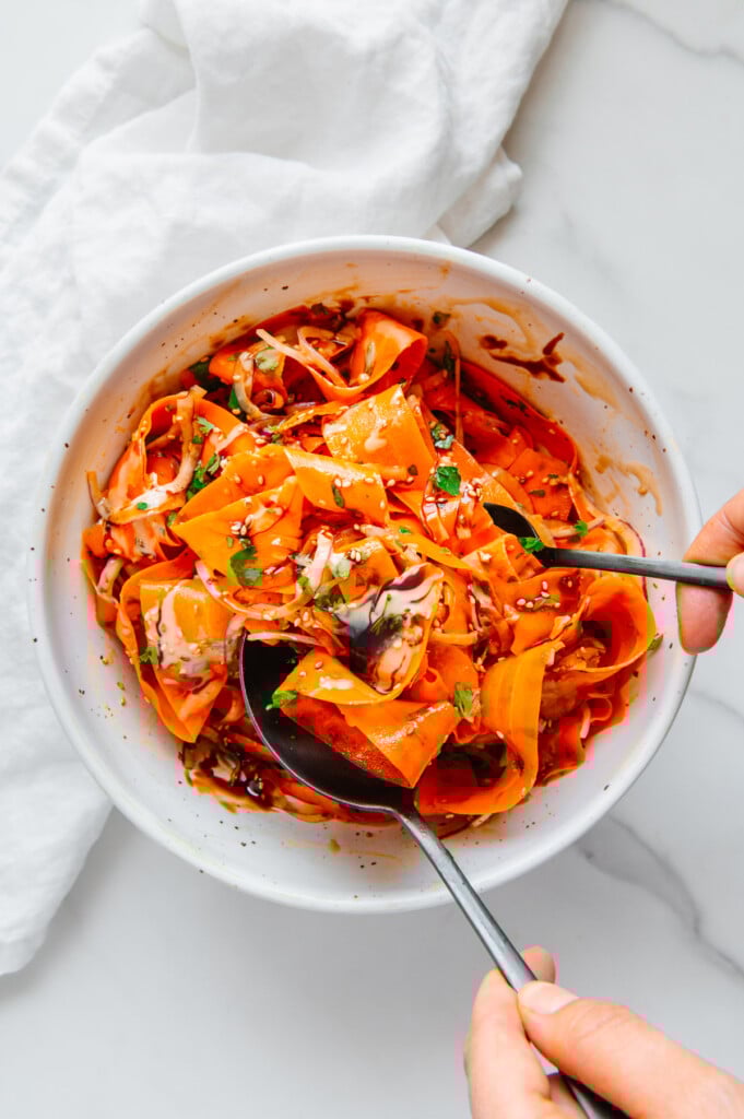 Two hands using utensils to toss ingredients of a carrot ribbon salad in a white bowl.