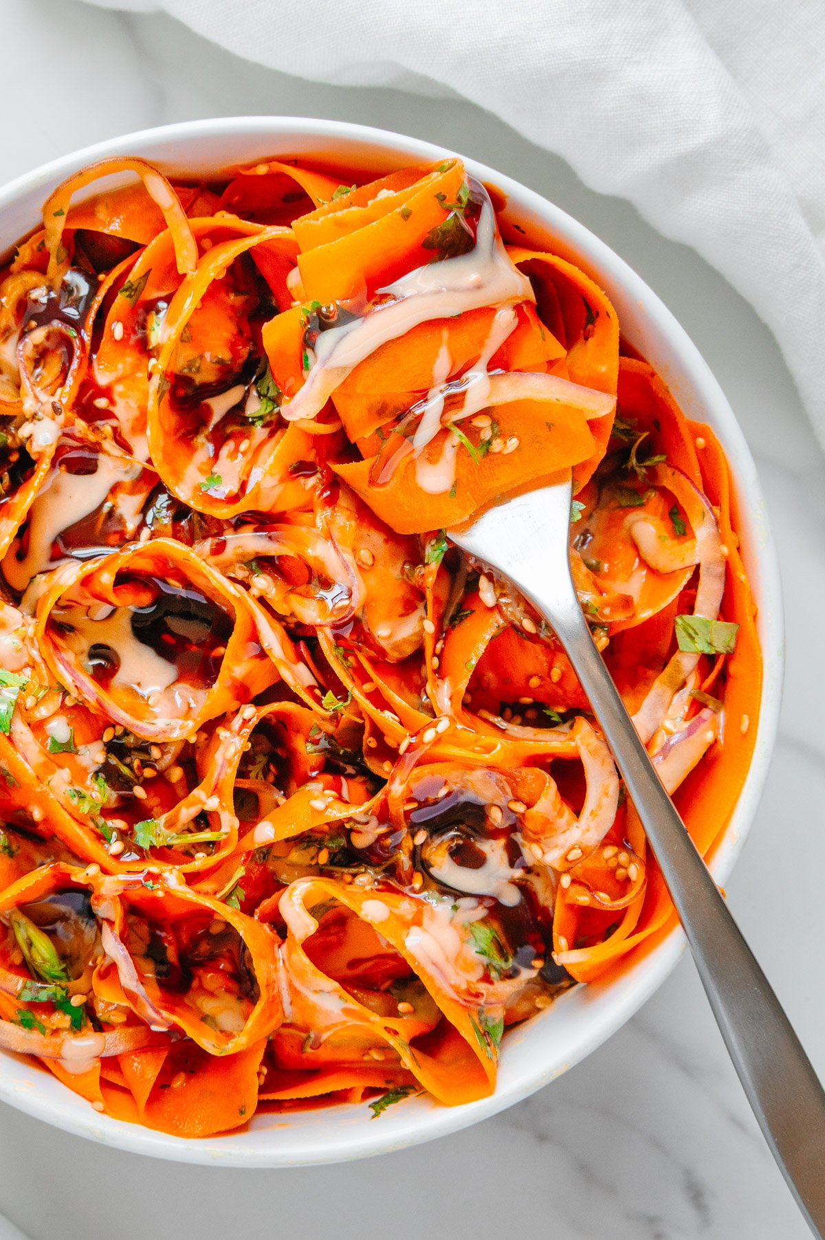 Carrot ribbon salad in a white bowl served with a silver fork with a carrot ribbon drizzled with tahini sauce wrapped around it.