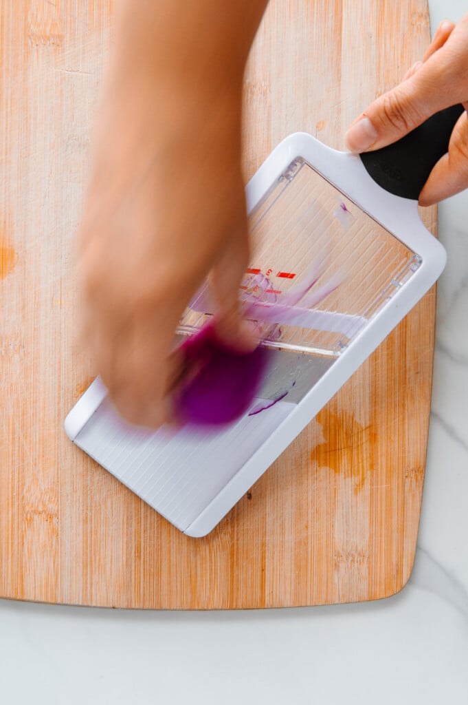 A hand using a mandoline to slice a red onion on a wooden cutting board.