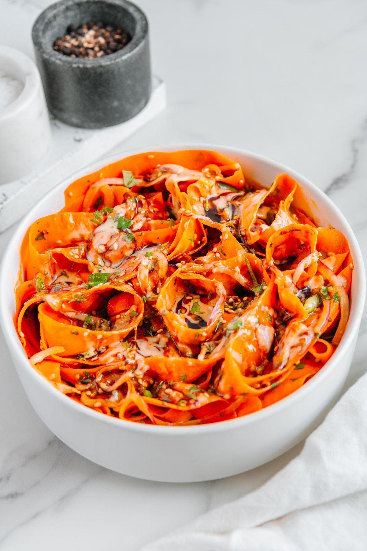 Carrot ribbon salad in a white bowl in front of salt and black pepper with a white marble background.