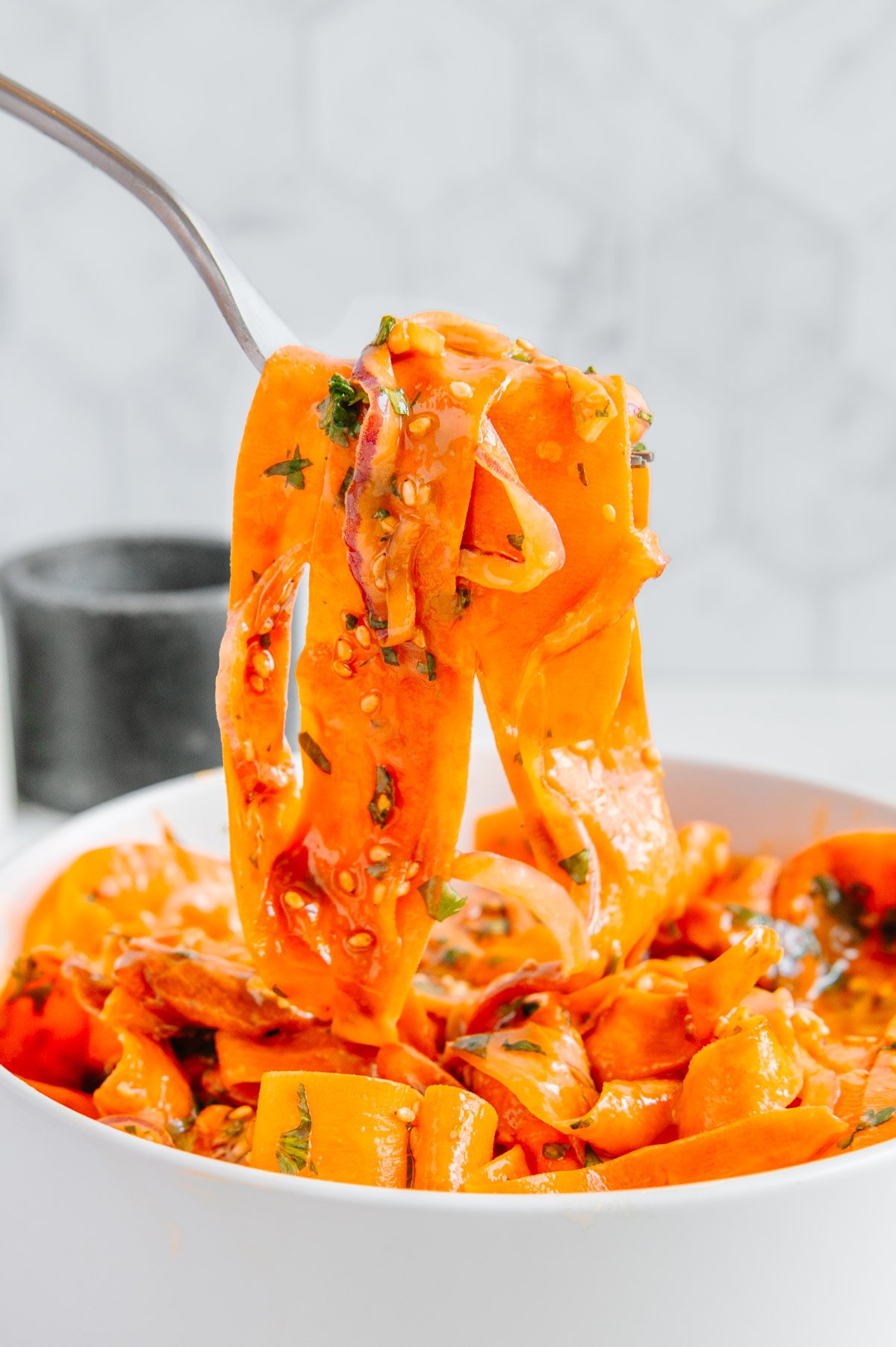 A fork lifting carrot ribbons up from a white bowl of carrot ribbon salad.