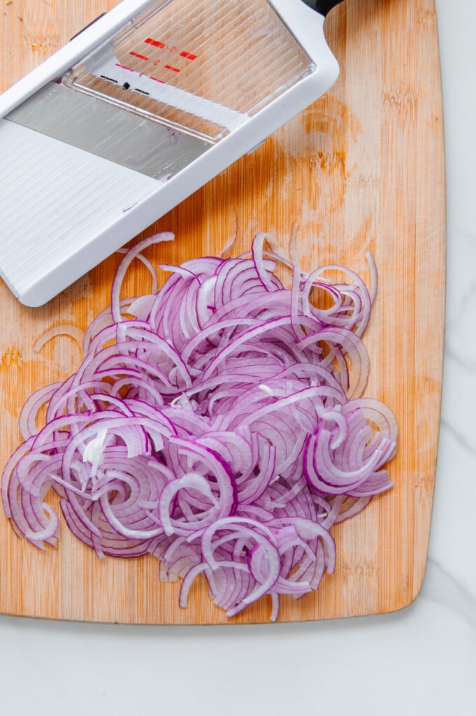 Thinly sliced onions on a wooden cutting board next to a mandoline.