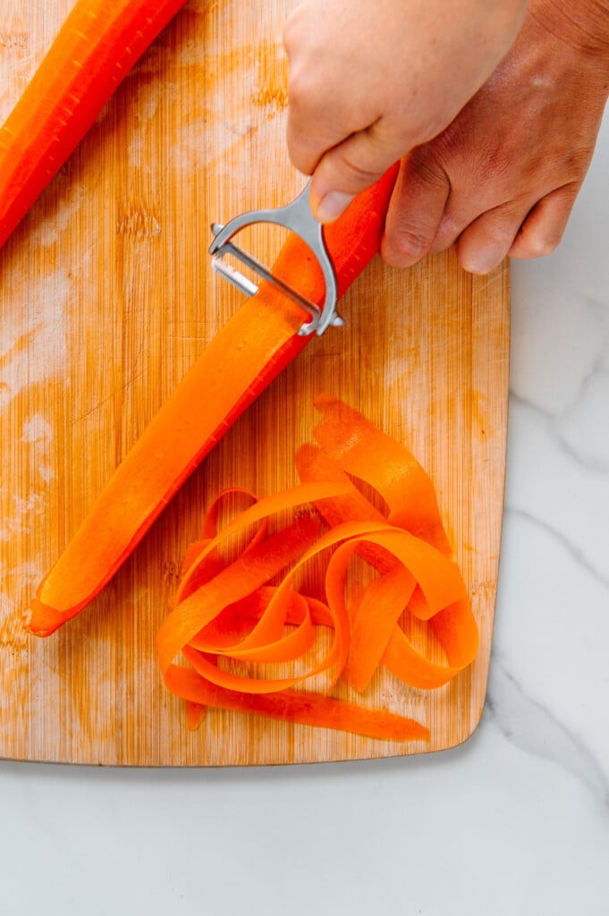 A hand using a vegetable peeler to peel carrot ribbons from a carrot on a wooden cutting board.