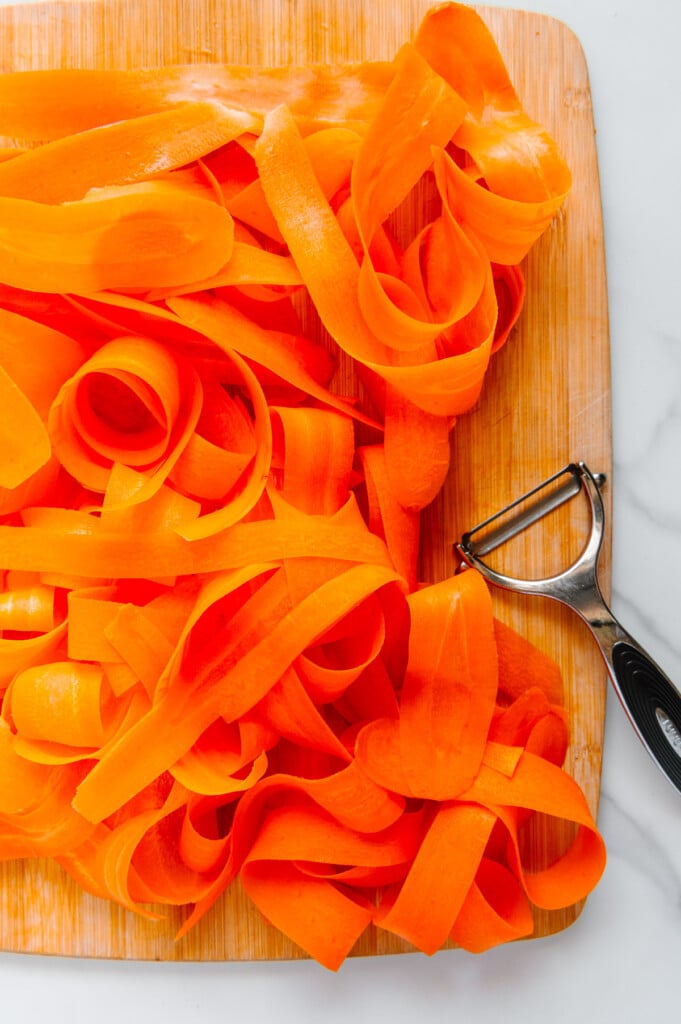 Carrot ribbons next to a vegetable peeler on a wooden cutting board.