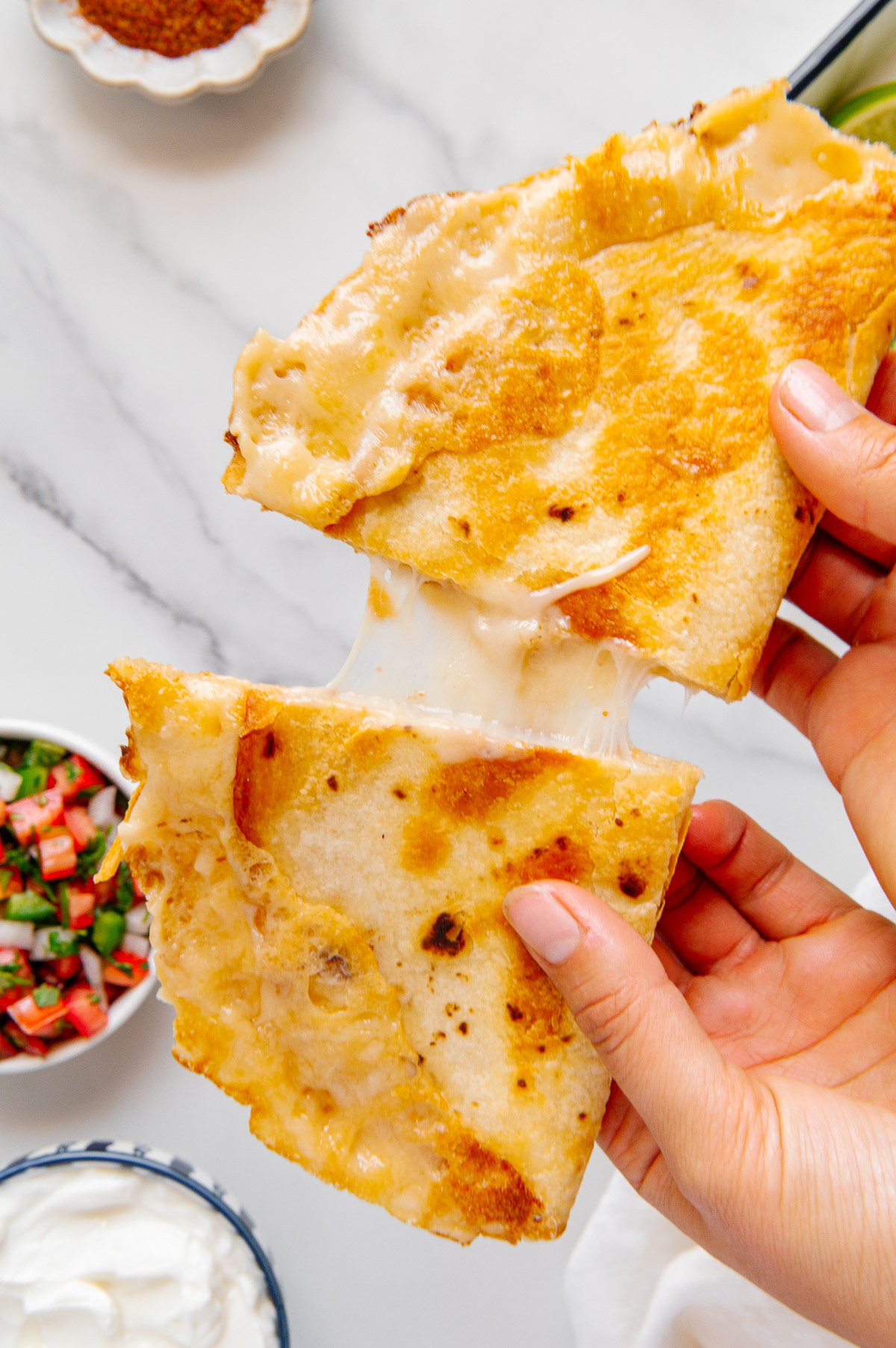 Hands pulling two halves of a vegan quesadilla apart above a white marble background.