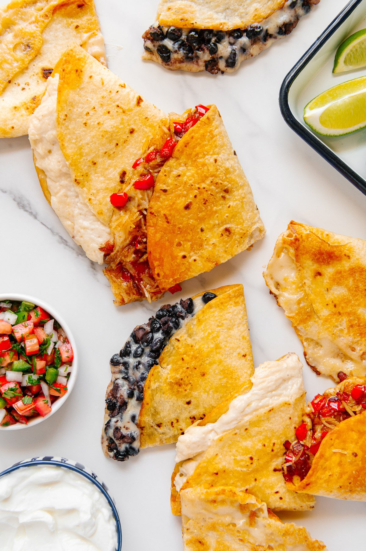 Various vegan quesadillas (one with veggies, another with black beans, etc.) alongside a bowls of pico de gallo and vegan sour cream with a white marble background.