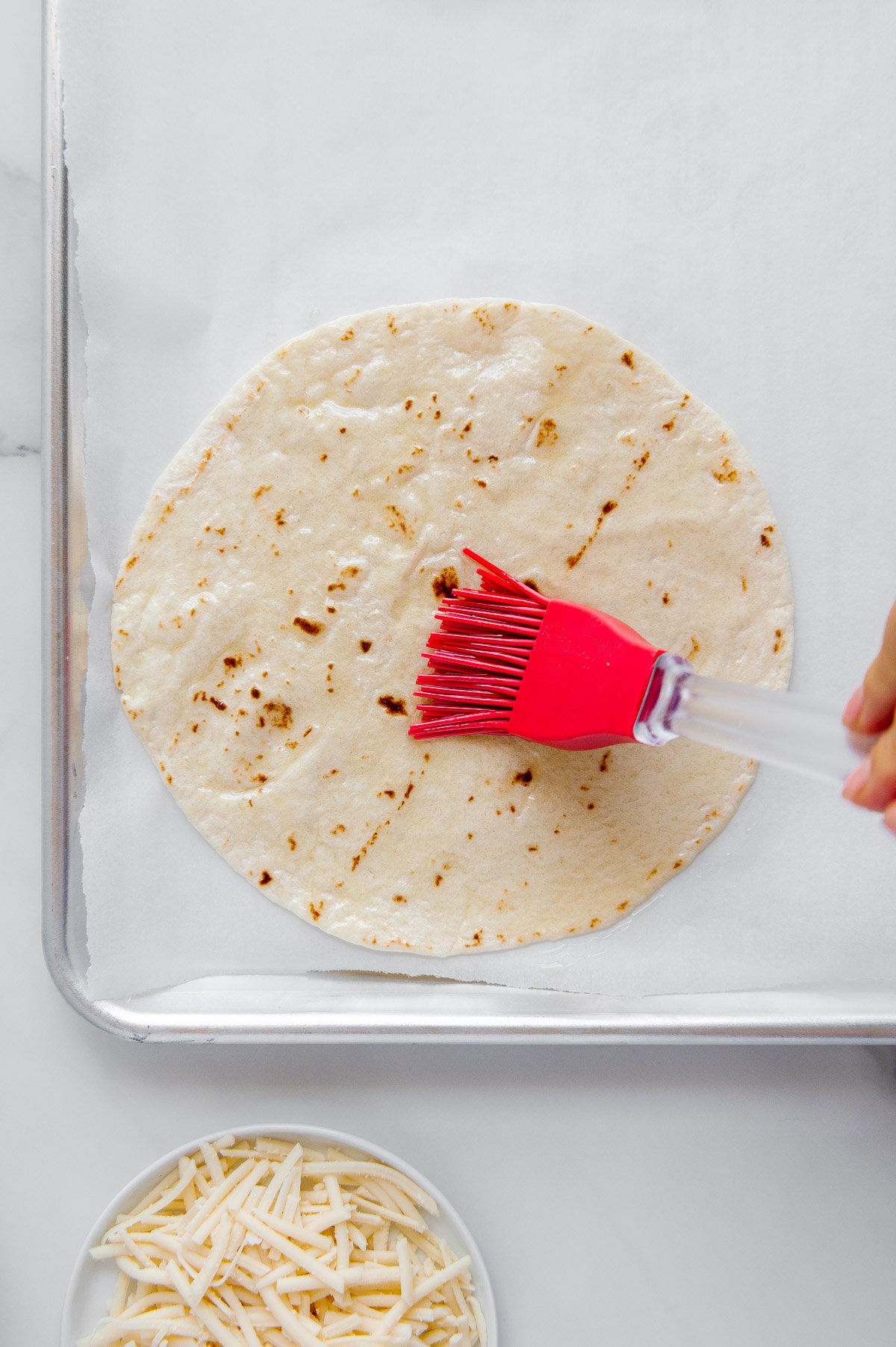 A hand using a red silicone brush to brush oil on a tortilla on a parchment paper lined sheetpan.