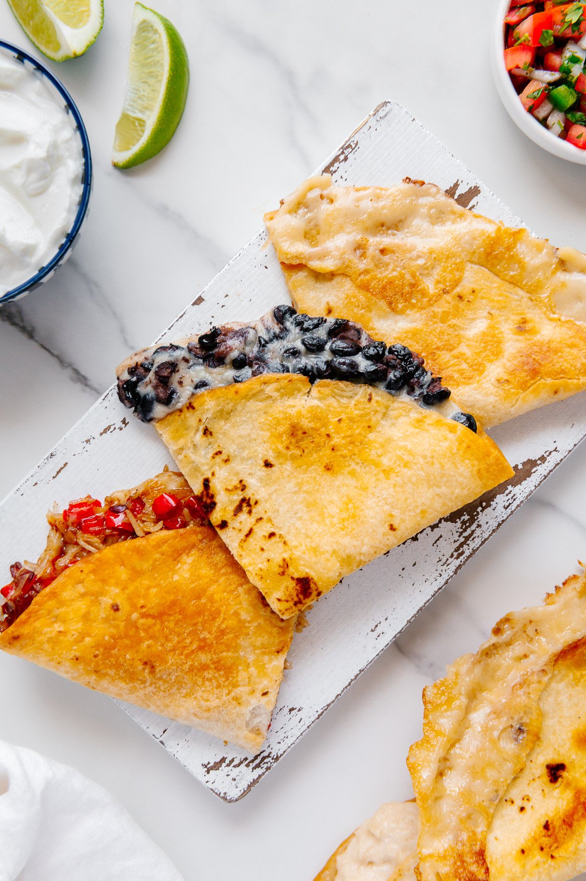 Three vegan quesadillas with various fillings on a white platter with a white marble background.