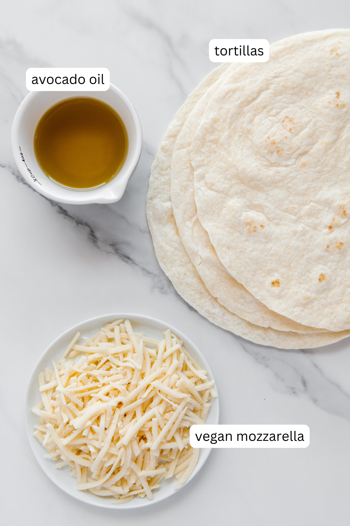 Tortillas, avocado oil and vegan shredded cheese with a white marble background.