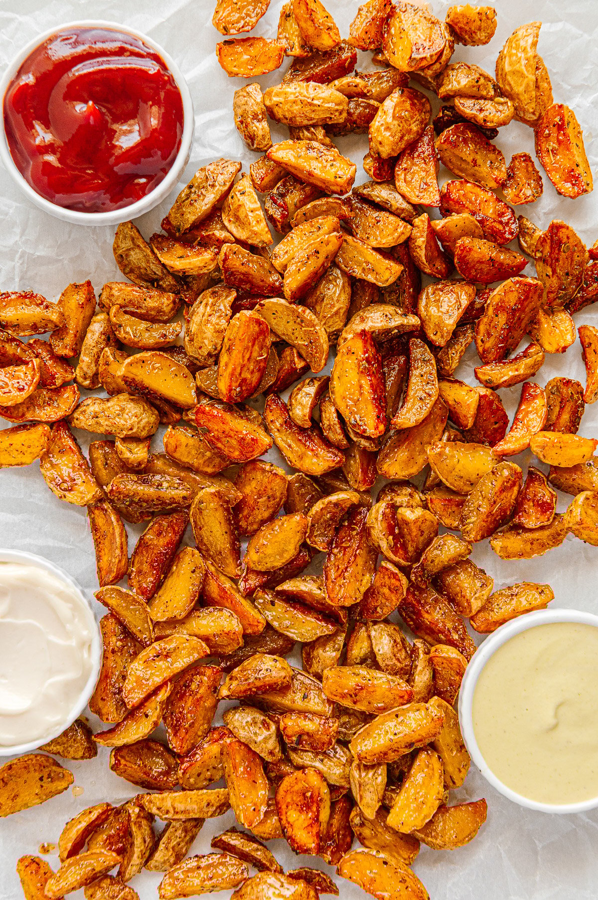 Crispy roasted potatoes on parchment paper served with ramekins of ketchup, honey mustard and vegan sour cream.