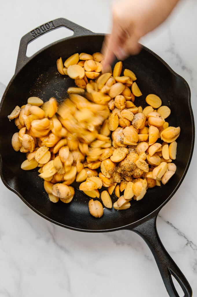 A hand sprinkling seasonings onto cut Baby Dutch Yellow potatoes in a cast iron skillet with a white marble background.