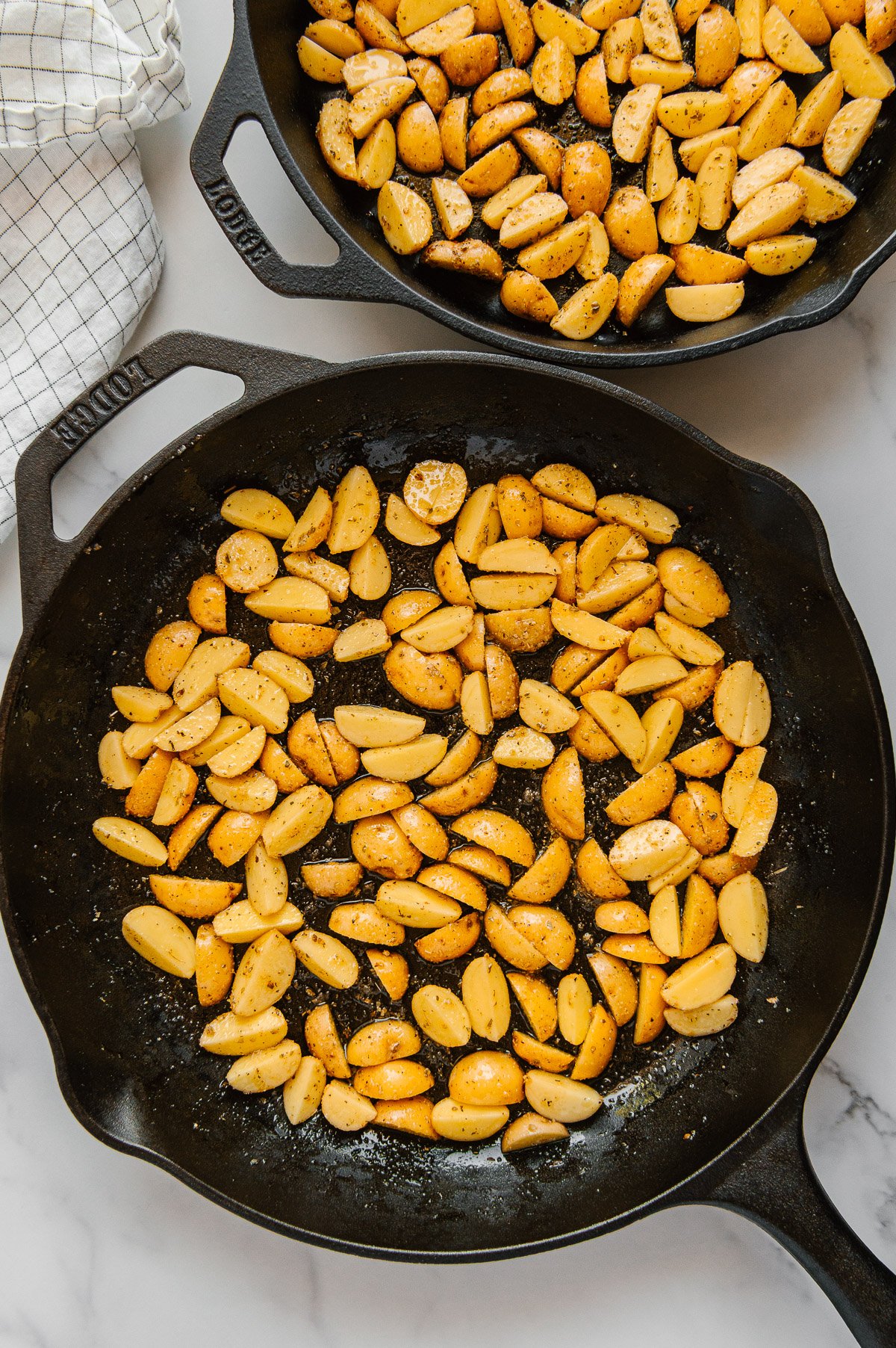 Sliced Baby Dutch Yellow potatoes in two cast iron skillets prior to roasting with a white marble background.