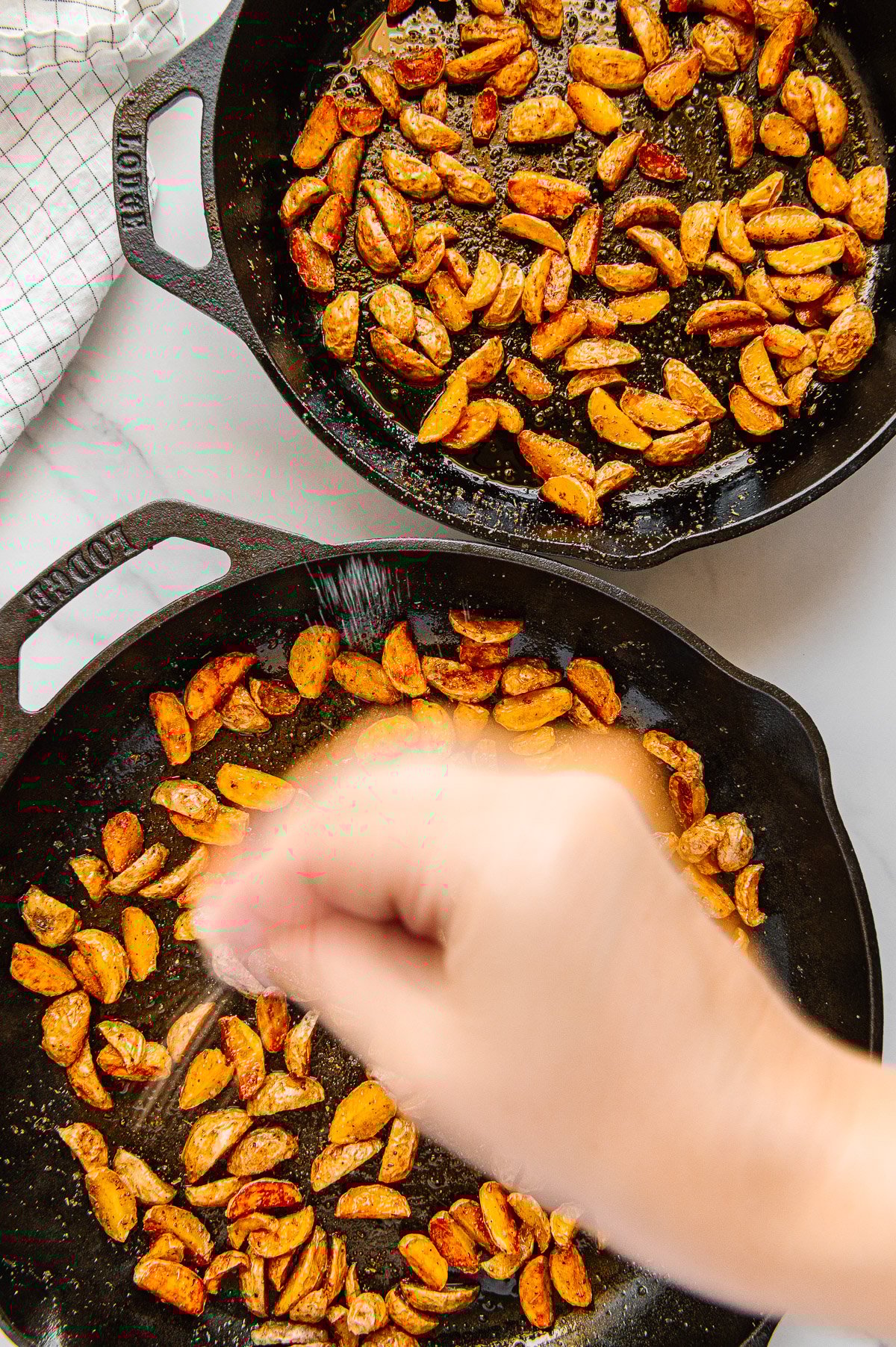 A hand sprinkling salt onto crispy roasted potatoes in a cast iron skillet.