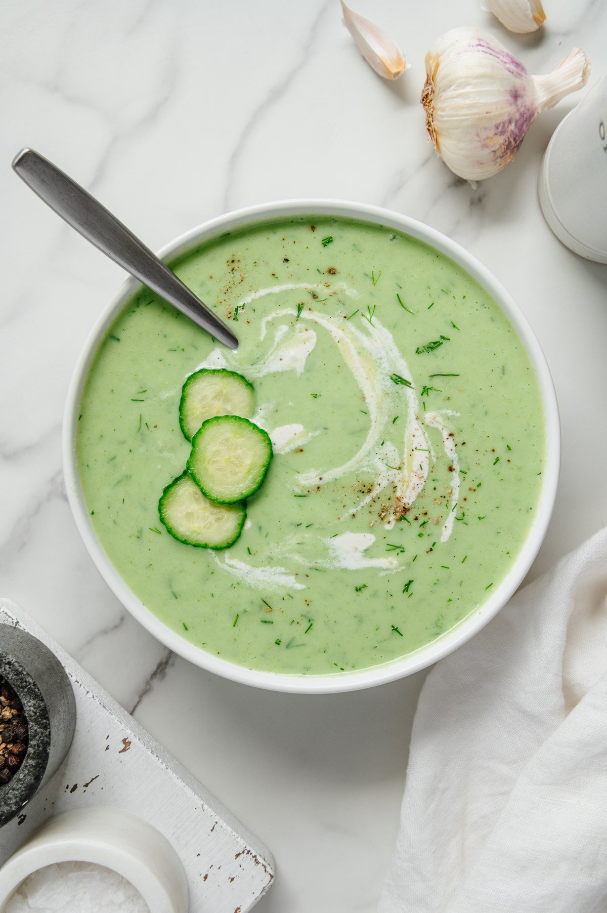 Cucumber soup garnished with cucumber, dill and vegan sour cream served in a white bowl with a spoon with a white marble background.