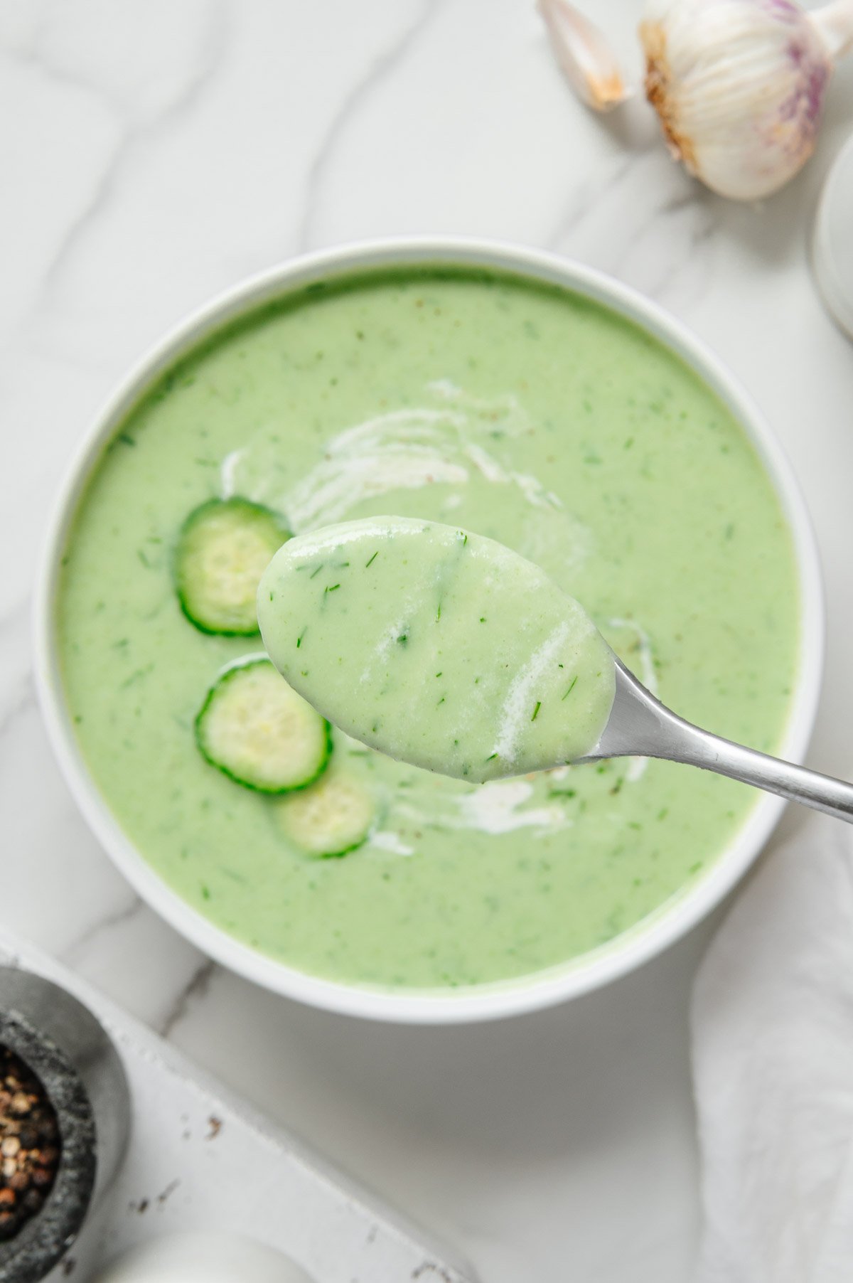 A close up shot of cucumber soup on a spoon held above a white bowl of cucumber soup.