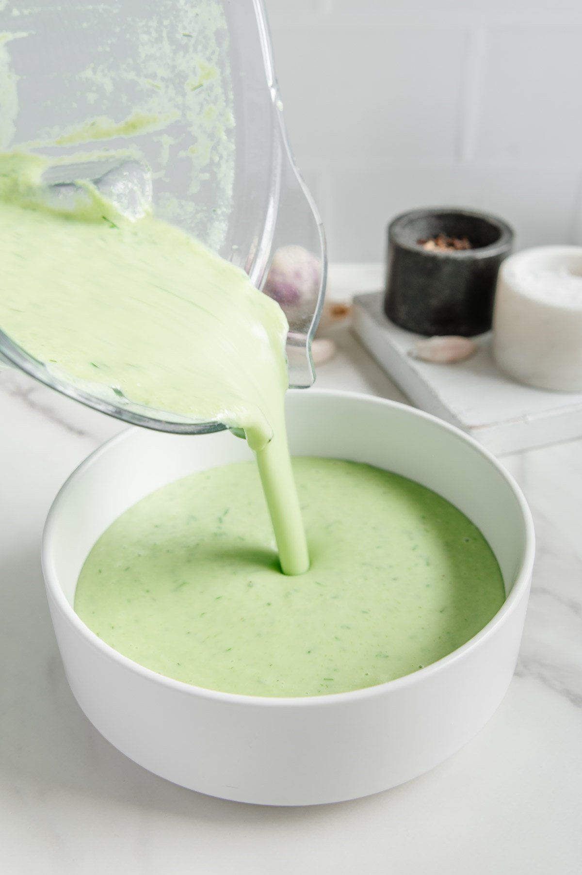 Cucumber soup being poured from a blender into a white bowl with a white marble background.