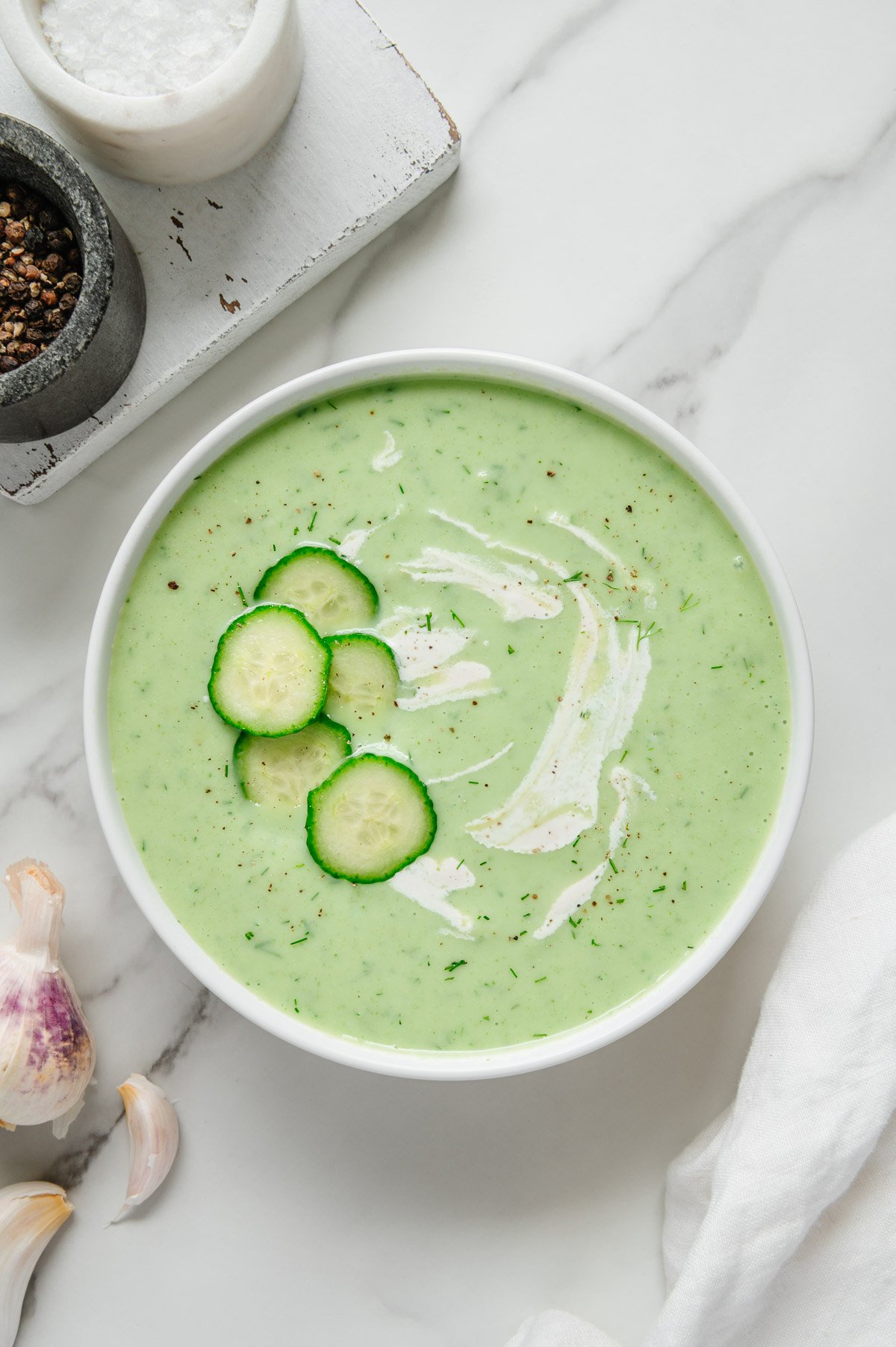 Cucumber soup garnished with sliced cucumber and vegan sour cream in a white bowl with a white marble background.