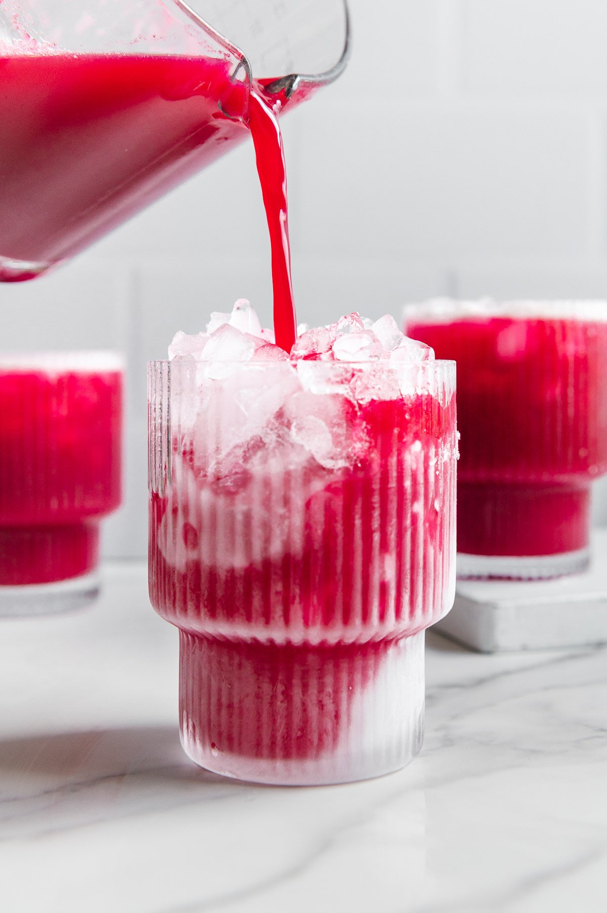 Fruit punch being poured from a blender into a glass filled with ice with a white marble background.