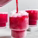 Fruit punch made with real fruit being poured from a blender into a glass with other glasses of fruit punch behind it with a white marble background.