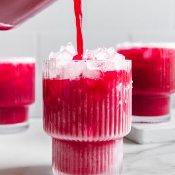 Fruit punch made with real fruit being poured from a blender into a glass with other glasses of fruit punch behind it with a white marble background.