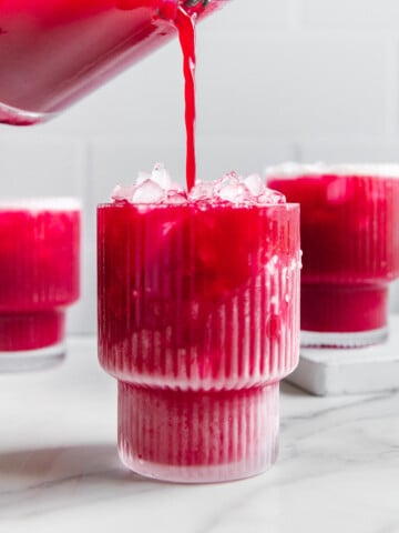 Fruit punch made with real fruit being poured from a blender into a glass with other glasses of fruit punch behind it with a white marble background.