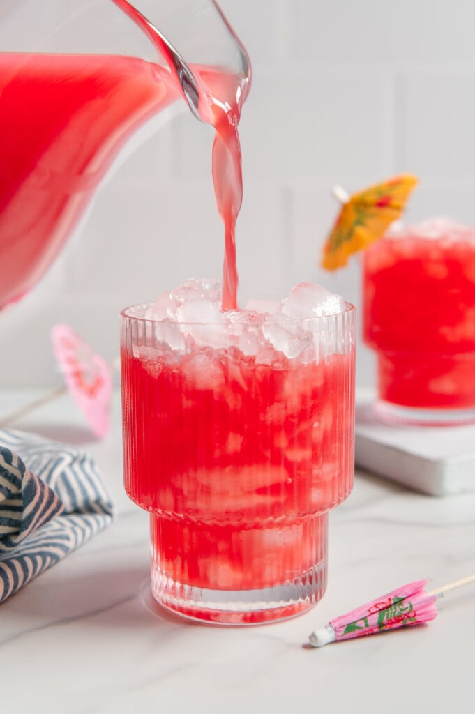 Hawaiian punch being poured from a glass pitcher into a glass of ice with a white marble background.