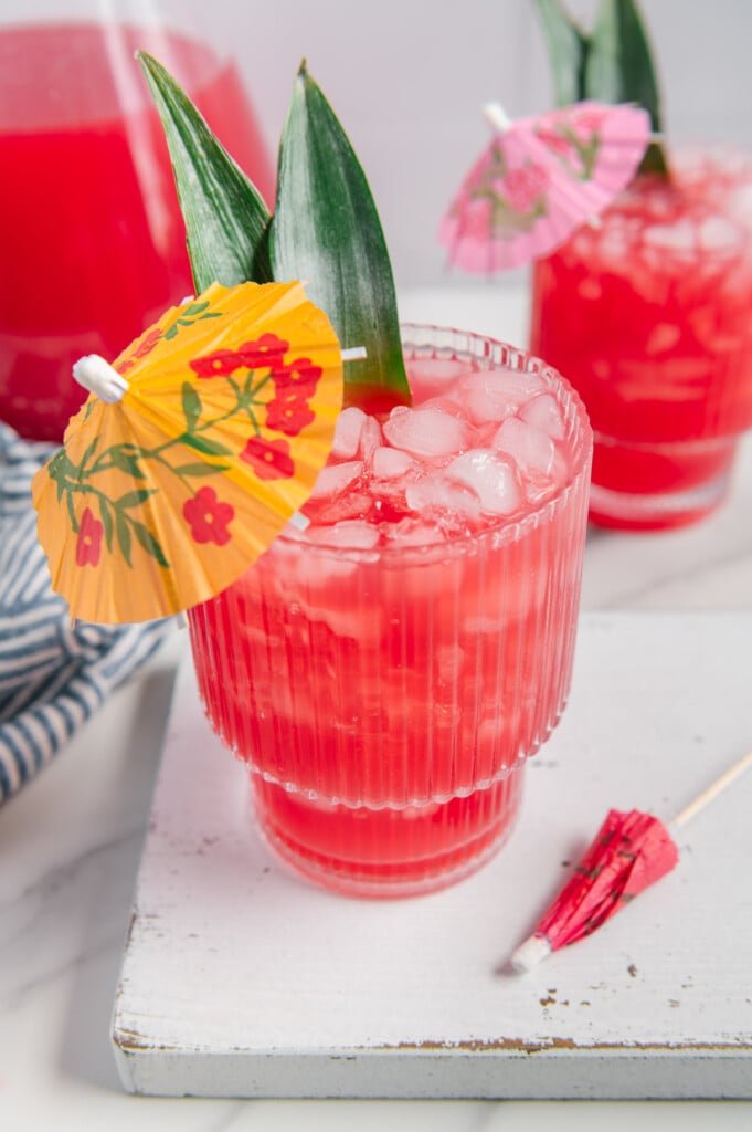 Hawaiian fruit punch in a glass served with ice, two pineapple leaves and a tropical yellow drink umbrella on a white cutting board.
