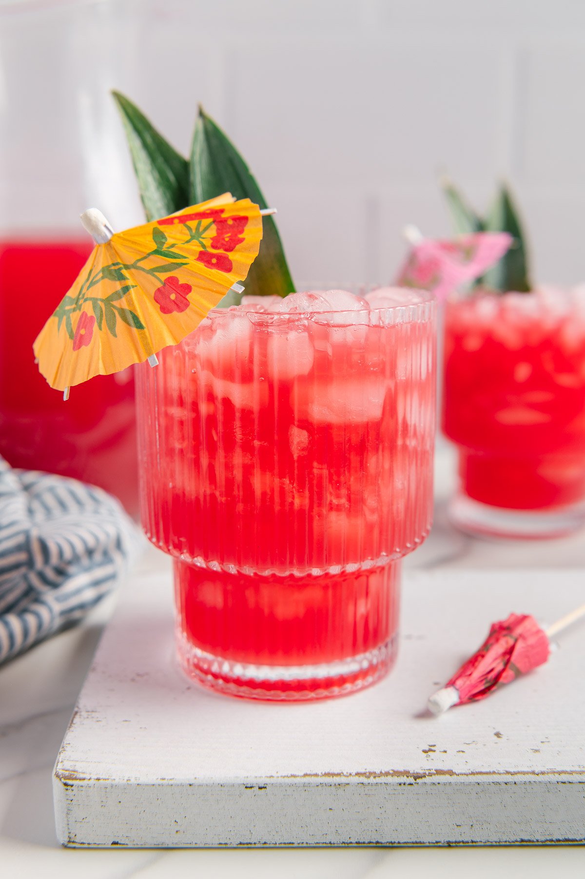 Hawaiian fruit punch in a glass served with pineapple leaves and a yellow tropical drink umbrella with a white background.