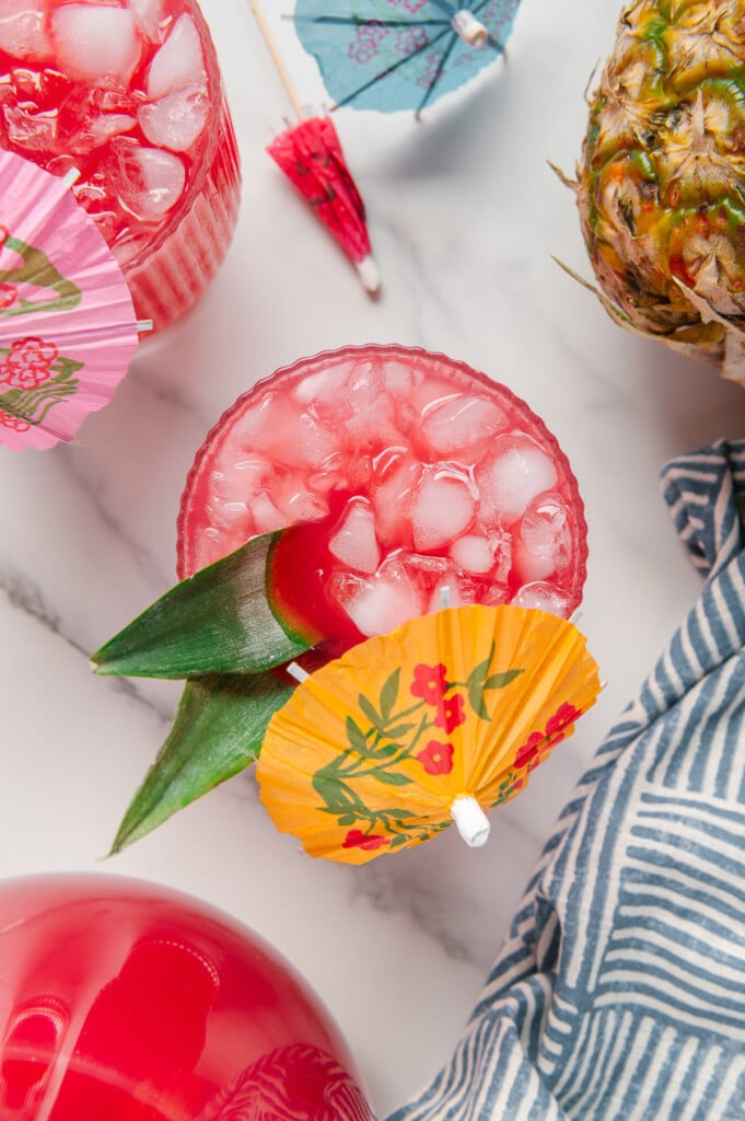 Overhead angle of Hawaiian fruit punch served with ice, pineapple leaves and a yellow tropical drink umbrella with a white marble background.
