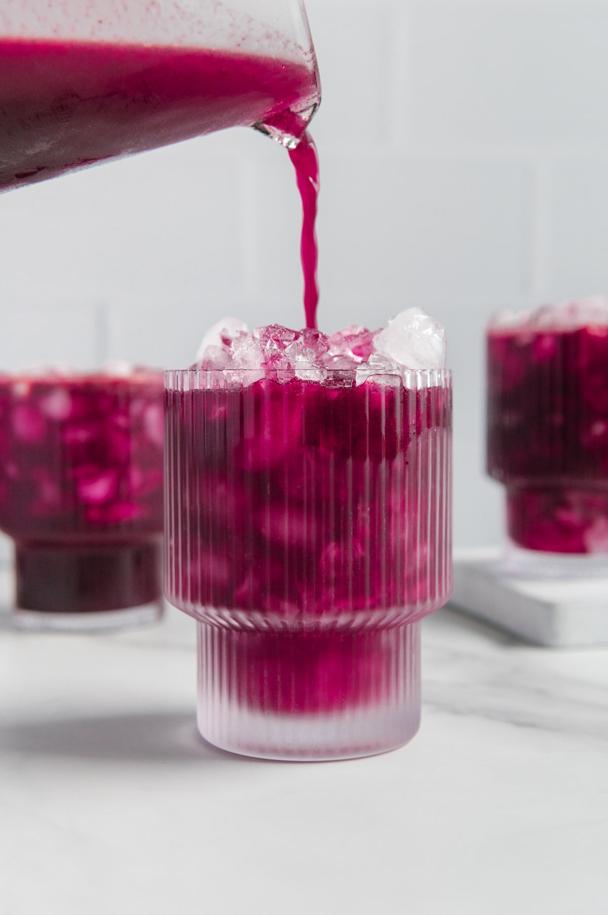 Pouring blueberry lemonade from a blender into a glass of ice with other glasses of blueberry lemonade in the background.