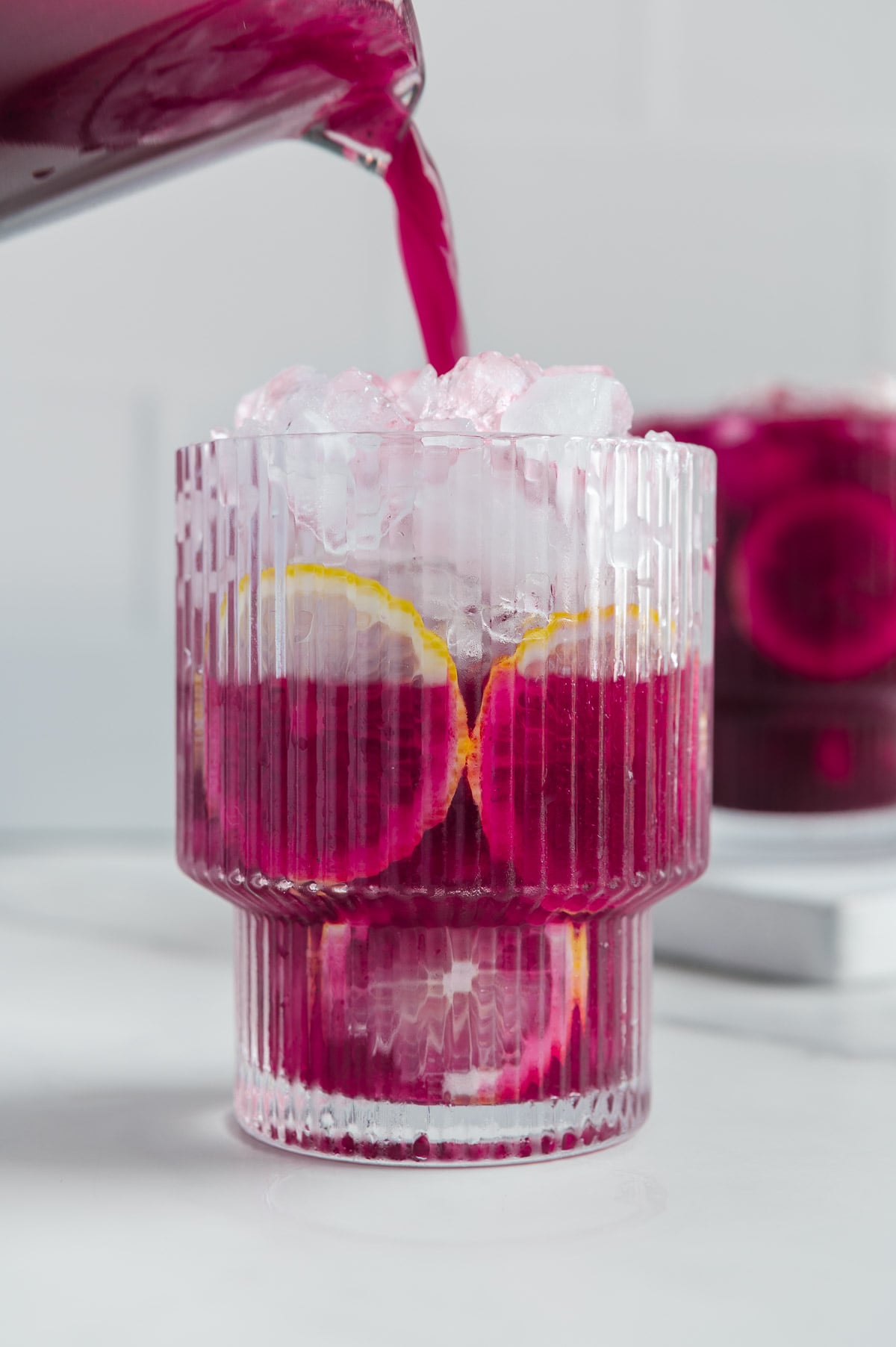 Blueberry Lemonade being poured into a glass full of ice with lemon inside on a white background