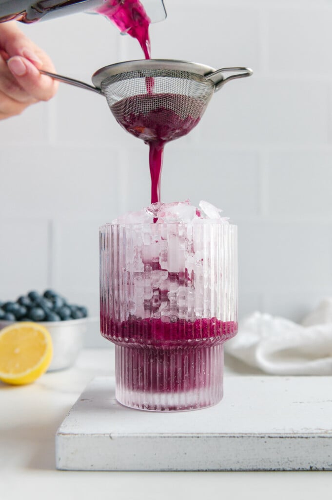 Using a strainer to pour blueberry lemonade from a blender into a glass of ice.