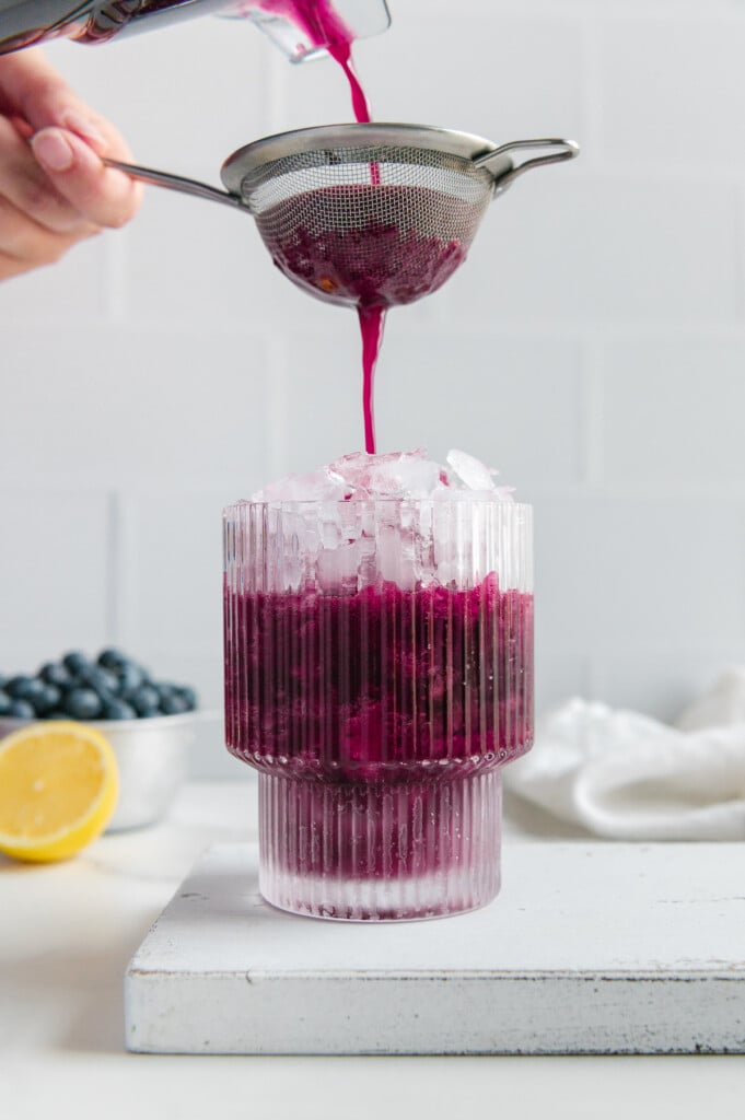 Using a strainer to pour blueberry lemonade from a blender into a glass of ice.