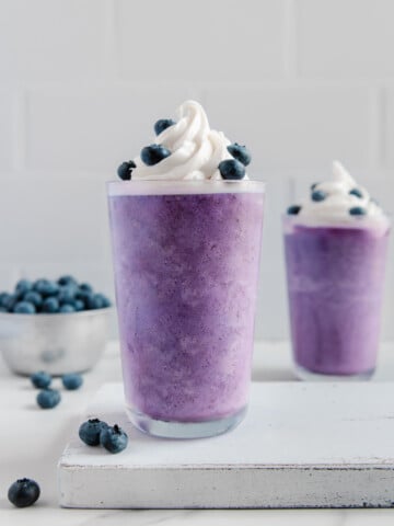 Two blueberry milkshakes topped with whipped cream and fresh blueberries with a bowl of blueberries in the background.
