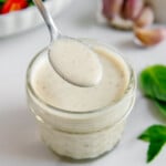 A spoonful of homemade vegan Italian dressing held over a jar of it with a basil, fresh garlic and a salad in the background.