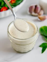 A spoonful of homemade vegan Italian dressing held over a jar of it with a basil, fresh garlic and a salad in the background.