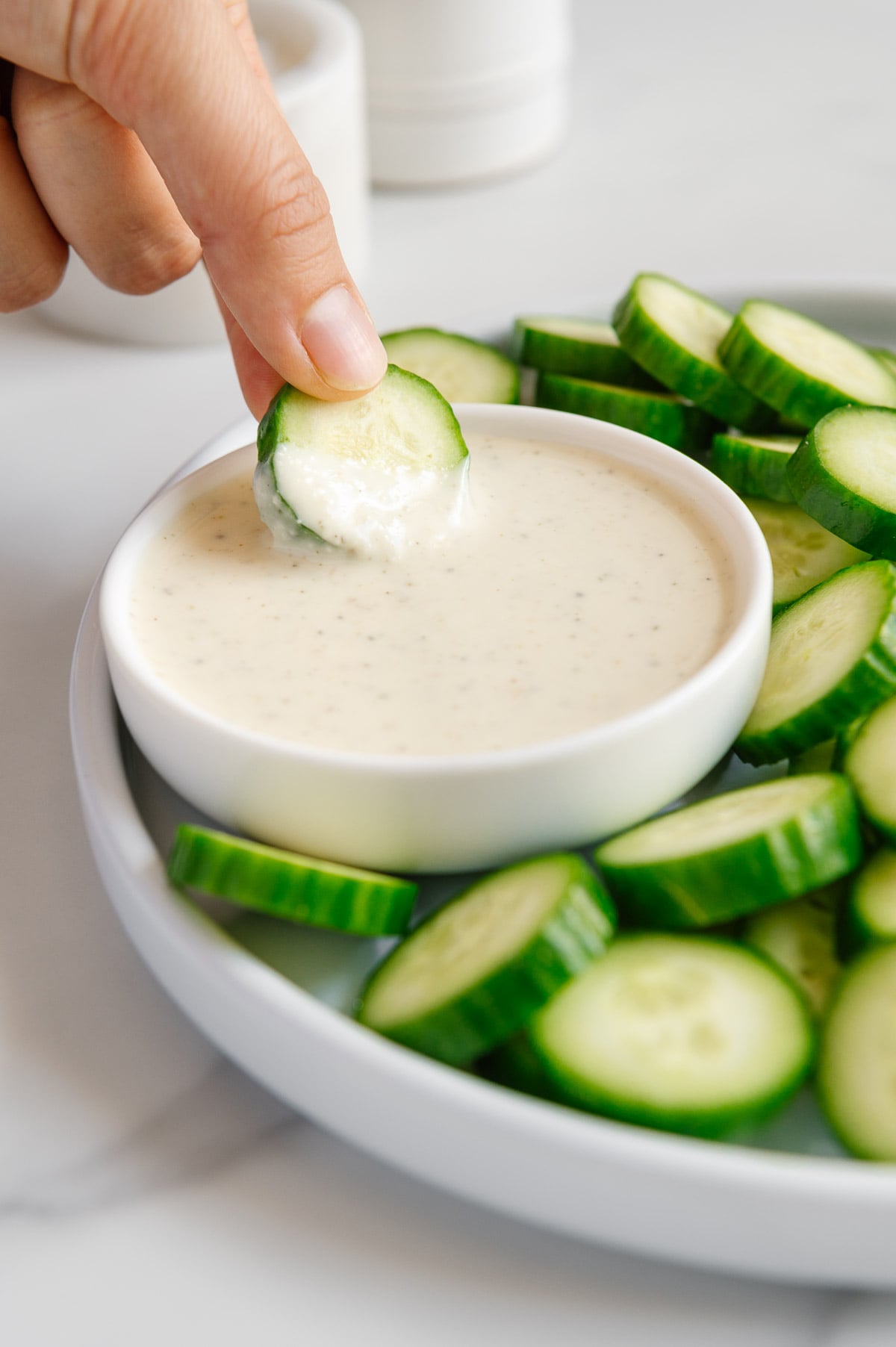 A hand dipping a sliced cucumber into a small white bowl of vegan Italian dressing with additional sliced cucumbers on a plate.