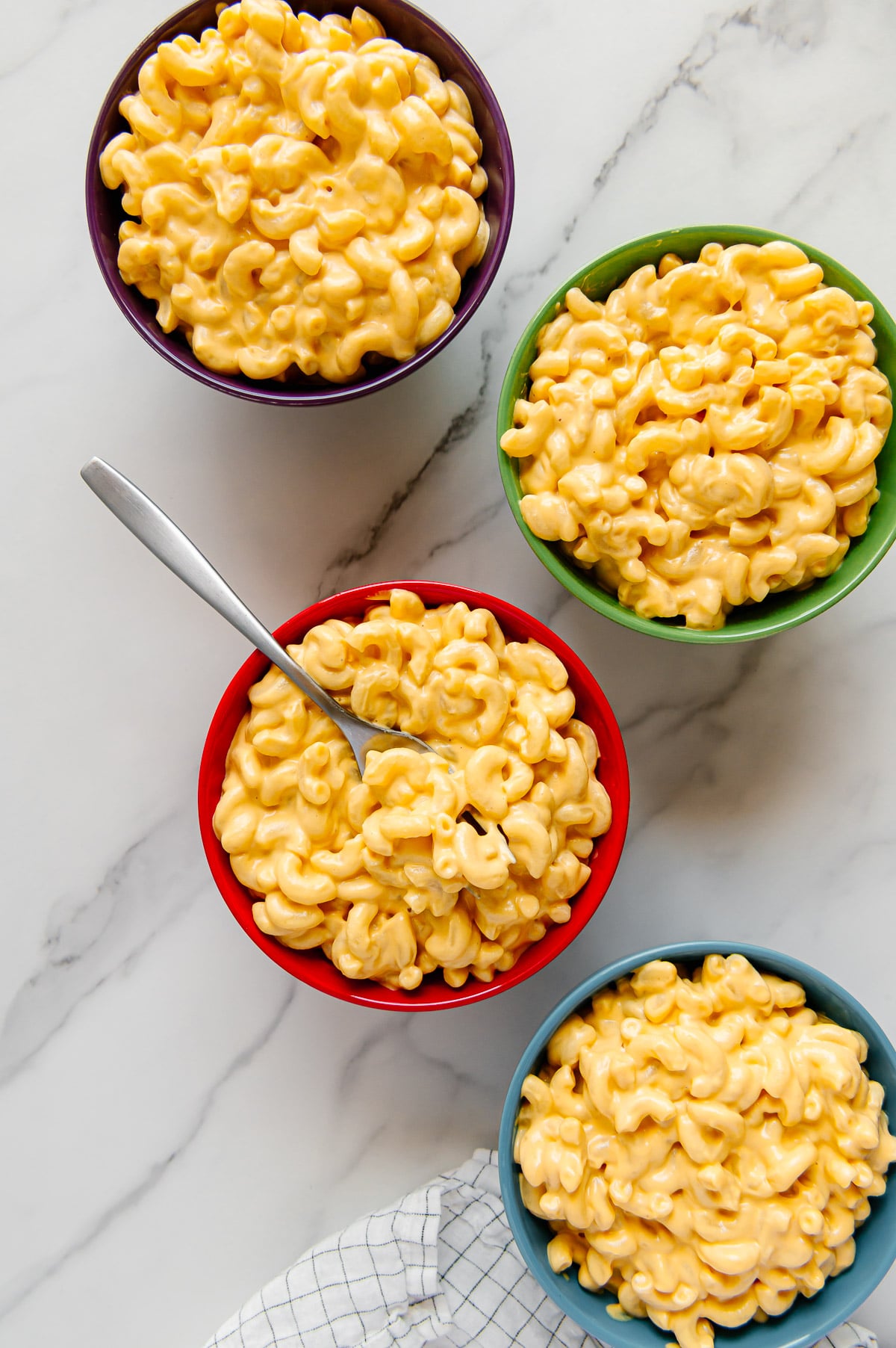 Vegan mac and cheese in four colored bowls (purple, green, red and blue) with a white marble background.