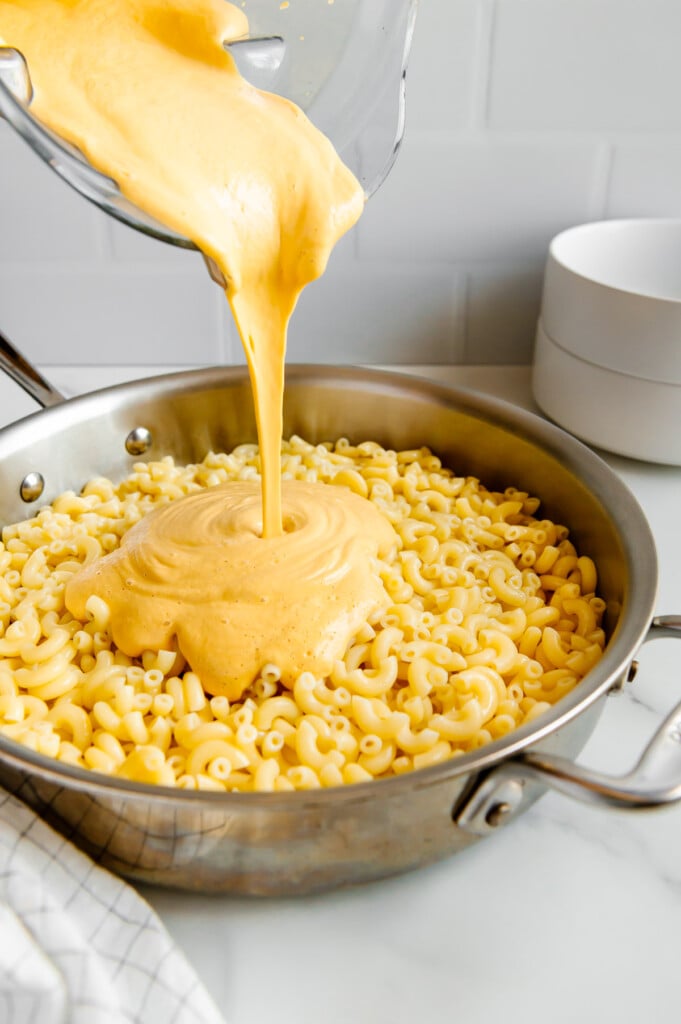 Pouring a vegan cheese sauce over elbow macaroni in a silver pot with a white background.