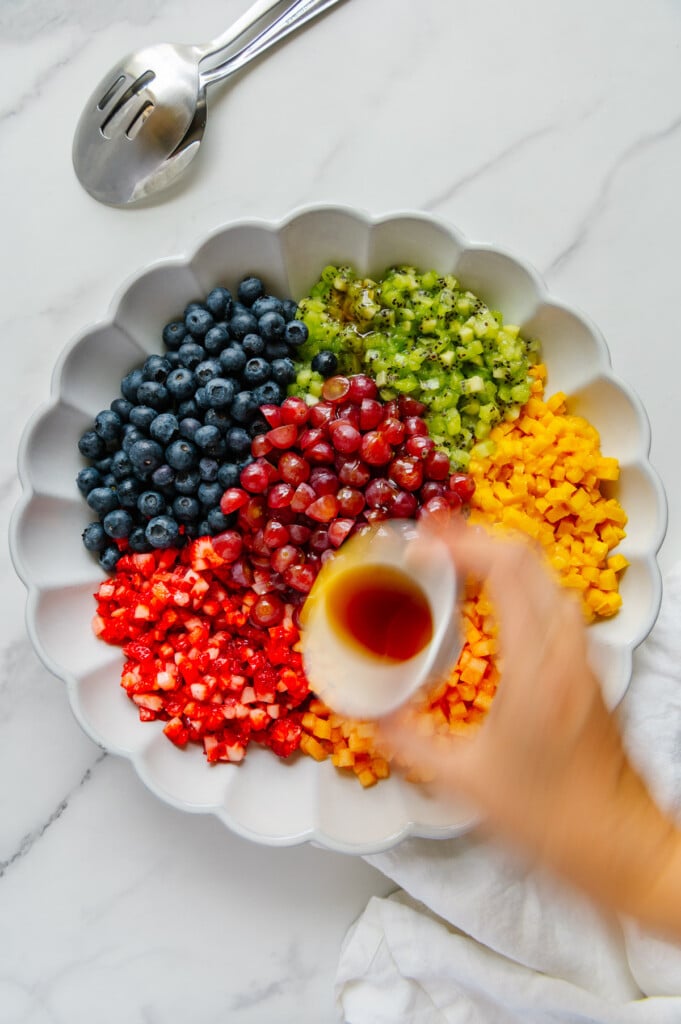 A hand pouring maple syrup onto a fruit salad in a white bowl prior to being tossed.