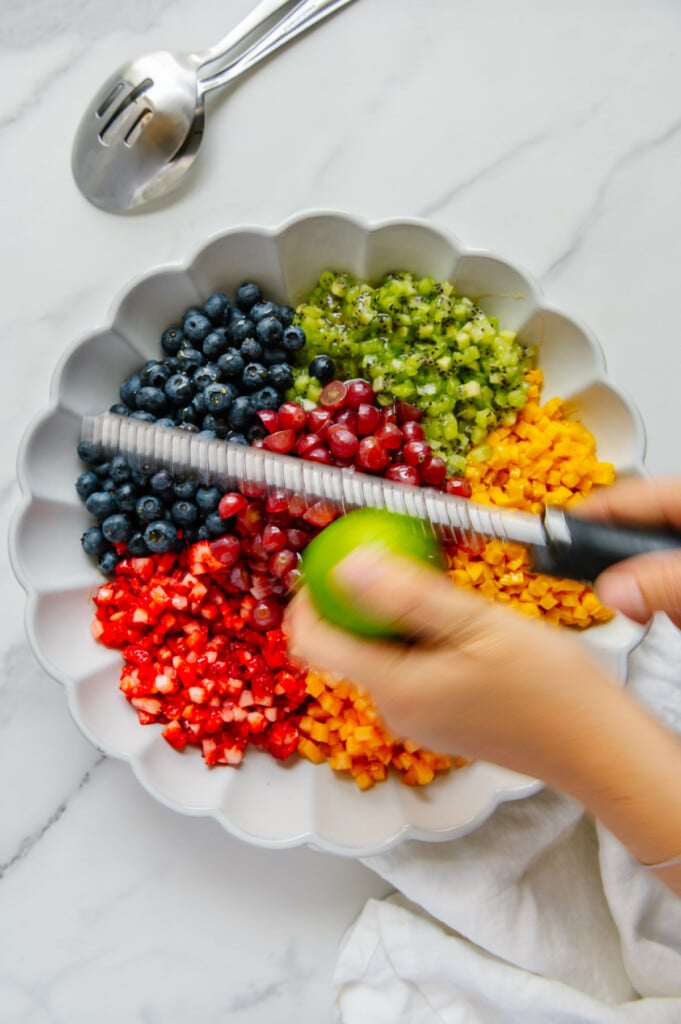 Hands zesting a lime over a fruit salad in a white bowl.