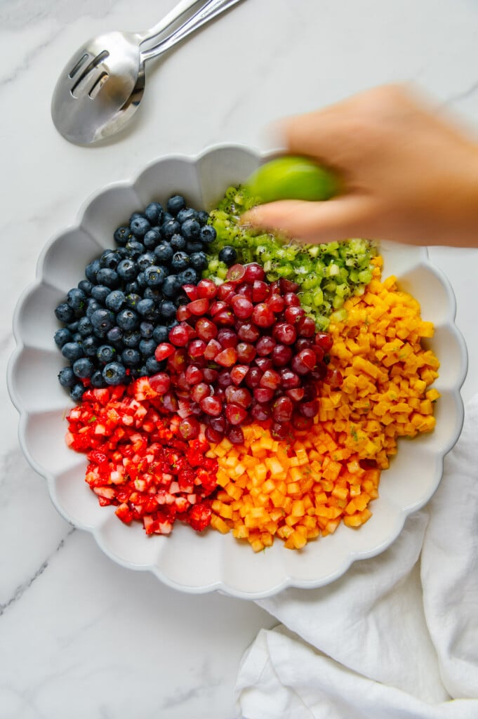 A hand squeezing a lime over a fruit salad in a white bowl.