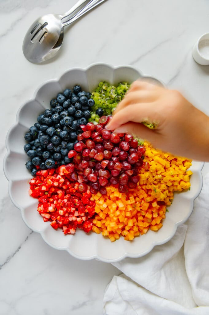 A hand adding a pinch of salt to a fruit salad in a white bowl.