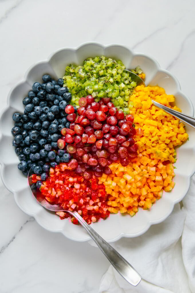Two spoons in a fruit salad in a white bowl prior to tossing.