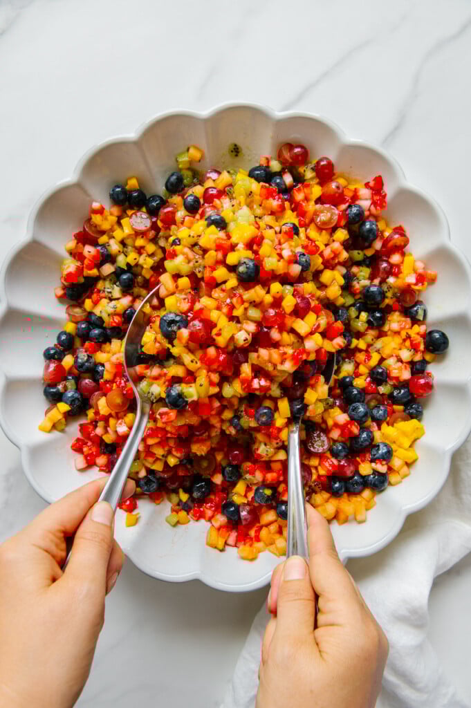 Two hands tossing a fruit salad using spoons in a white bowl.