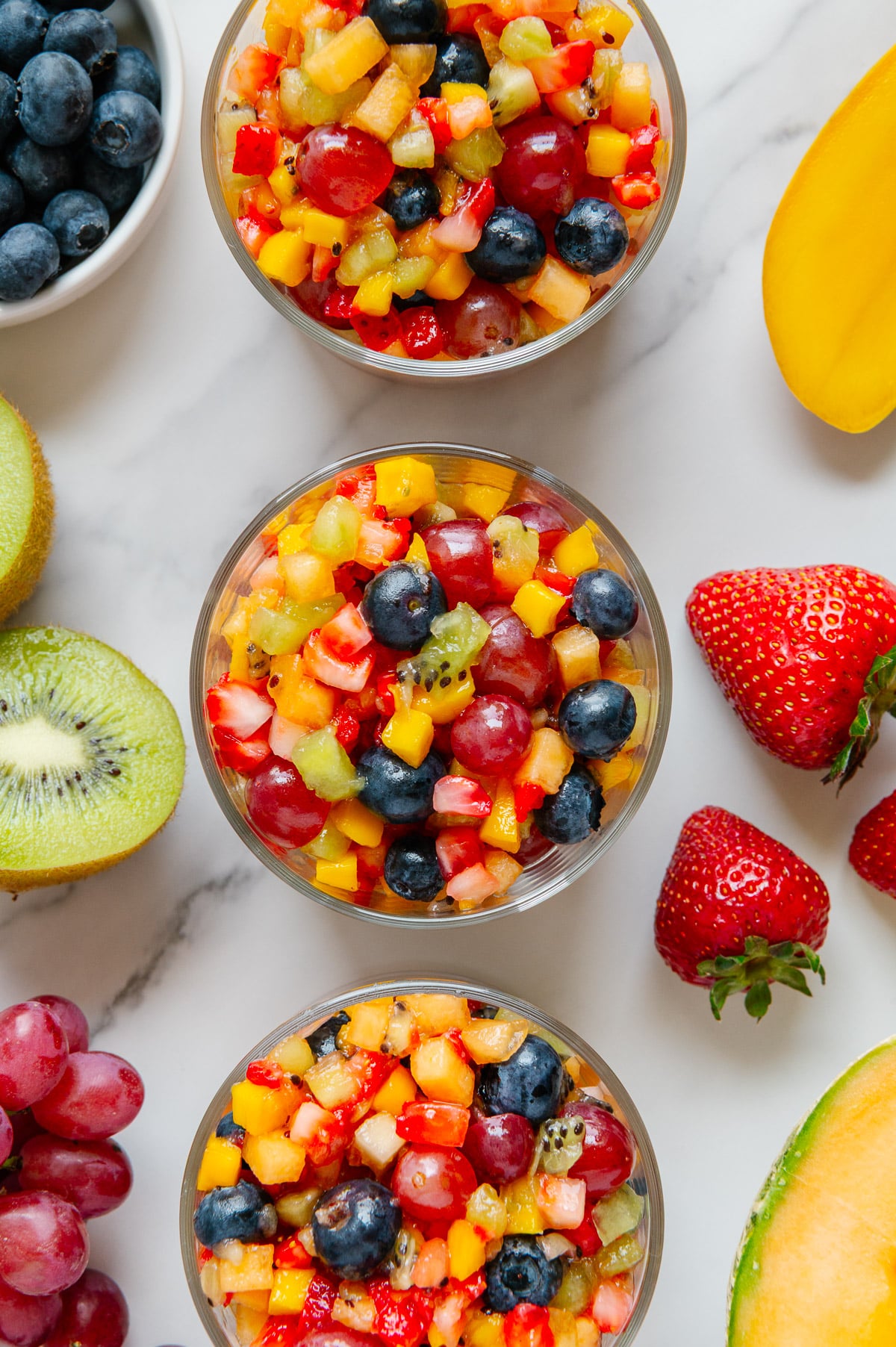 Nature's Skittles fruit salad in small glass bowls alongside fresh fruit with a white marble background.