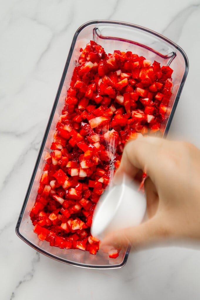 A hand pouring salt over finely chopped strawberries in a vegetable chopper.