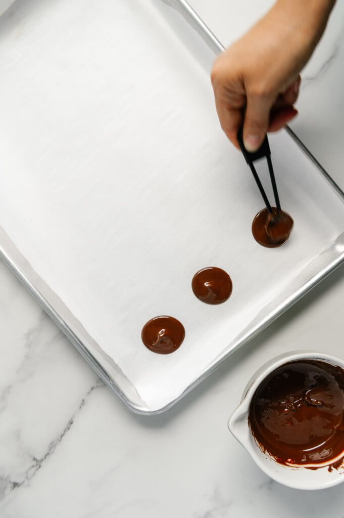 A hand using a teaspoon to create small chocolate discs on a parchment-lined baking sheet.